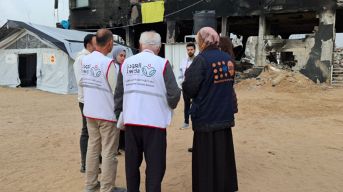 Des agent·e·s de santé et du personnel de l’UNFPA se tiennent devant un immense bâtiment détruit à côté d’une tente médicale portant le logo de l’UNFPA